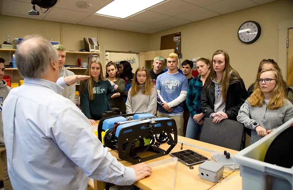 A group of high school students gathered around a professor as he explains a marine science tool produced by U N E students